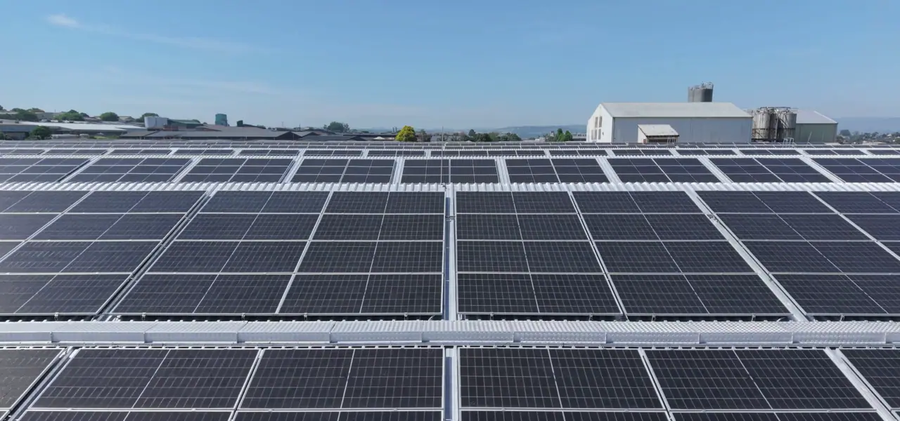 Low aerial view across rows of rooftop solar modules on the Belgotex factory, with a white plant building and silos on the skyline