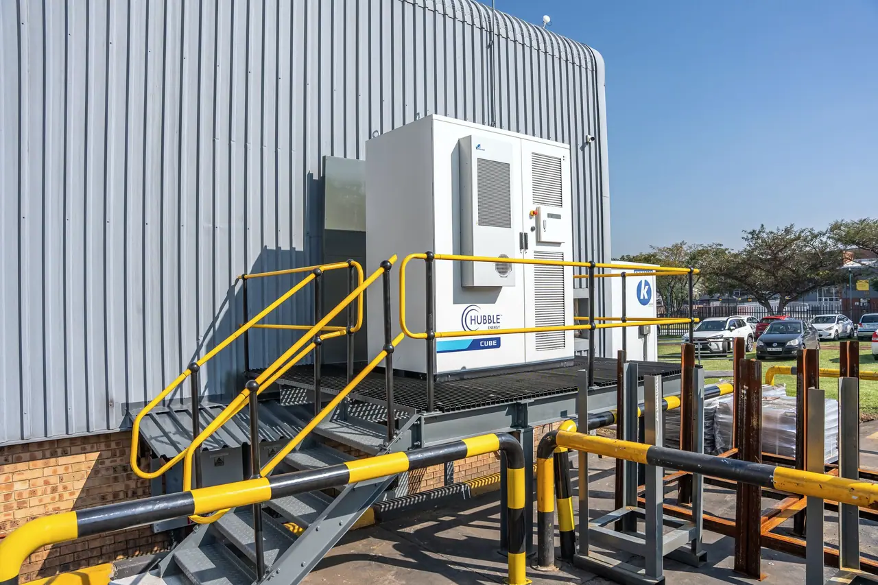 A battery storage cabinet on a raised platform with yellow safety railings, beside a factory wall
