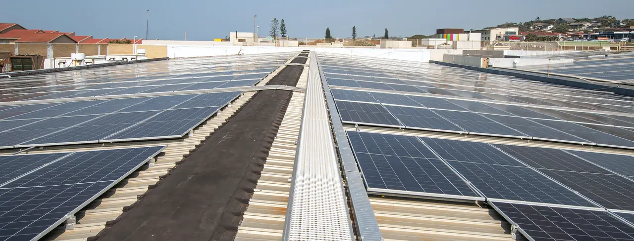 A steel walkway running between long rows of solar modules on the Shelly Centre roof, with the town beyond