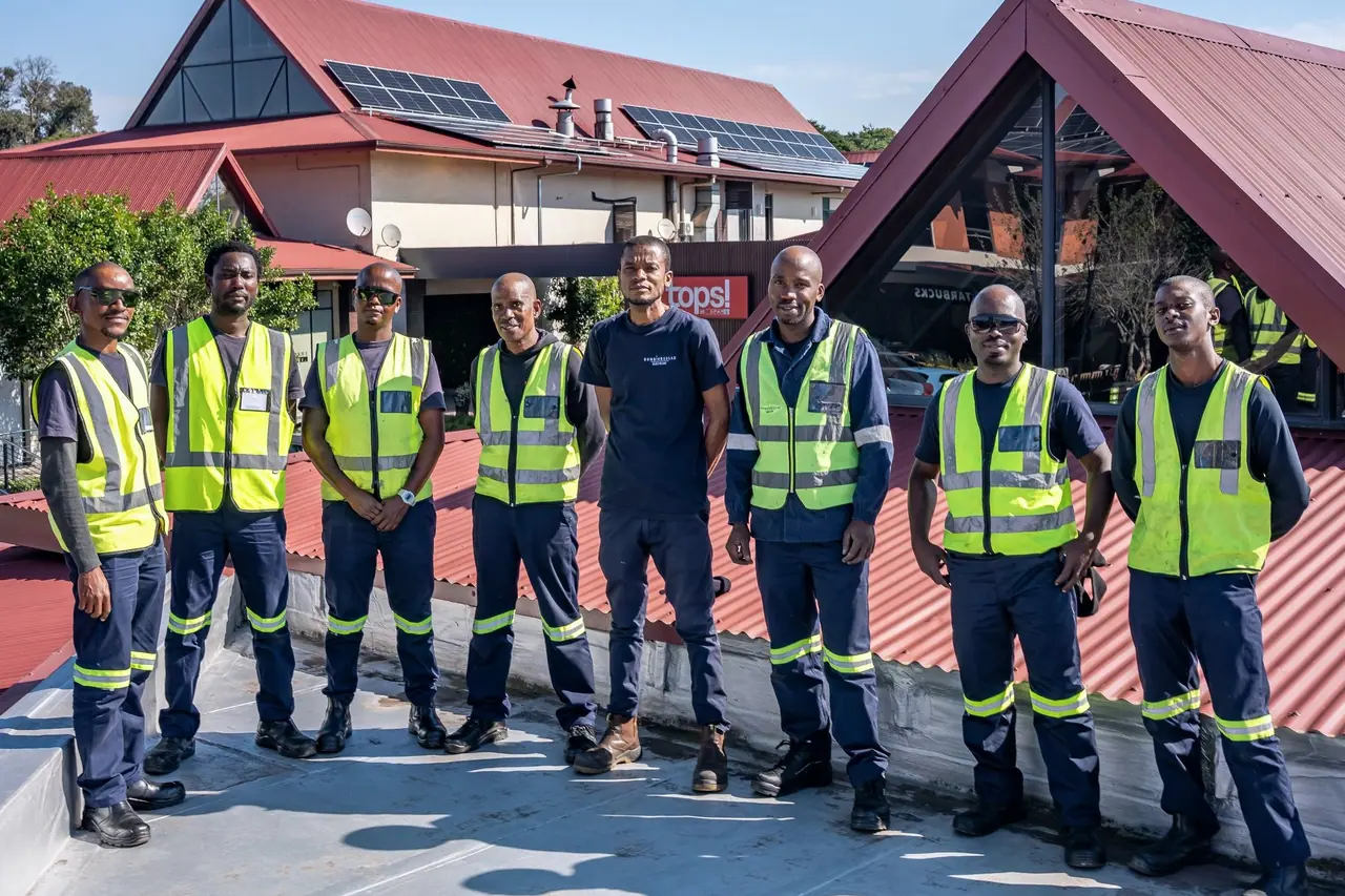 Eight members of the Sunbird site team in work wear and high-visibility vests on a flat roof at Lillies Quarters, with rooftop solar modules on the building behind them